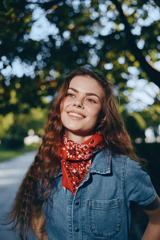smiling woman outdoors in denim jacket and red scarf, natural light, green blurred background, casual style, warm sunlight, happy expression