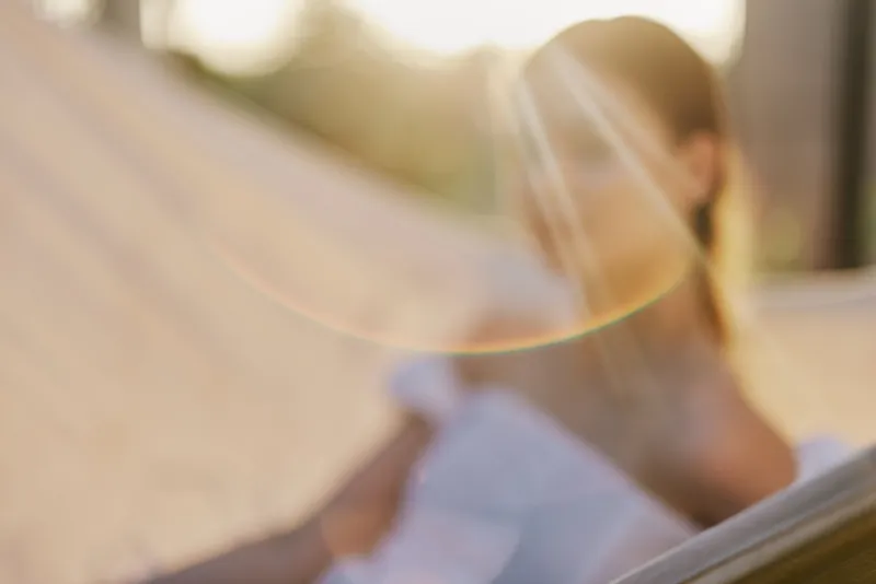 Young woman relaxing in a hammock with soft sunlight, wearing a white dress, embodying tranquility and summer vibes in a nature setting