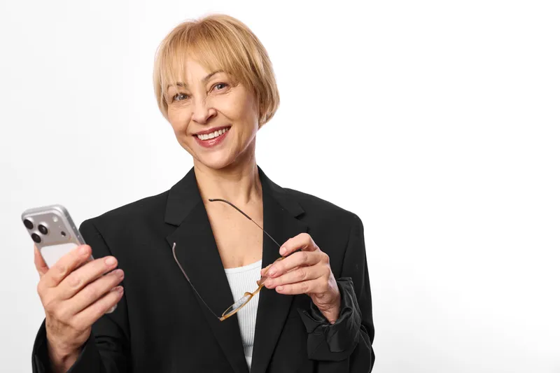 Woman senior smiling holds a smartphone and gestures in a business portrait, conveying technology and communication on a clean white background for digital training