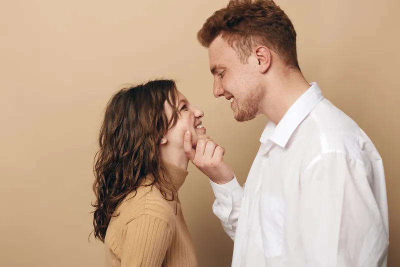 Smiling young couple in casual clothes share a tender moment, man gently touching woman’s cheek. Warm beige background creates intimate and loving atmosphere, concept of romance and connection.