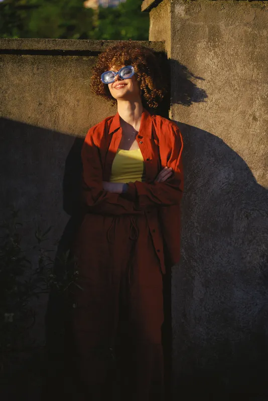Cheerful woman outdoors in sunglasses, bright orange coat and yellow top, leaning against a sunlit wall, carefree lifestyle moment with warm tones