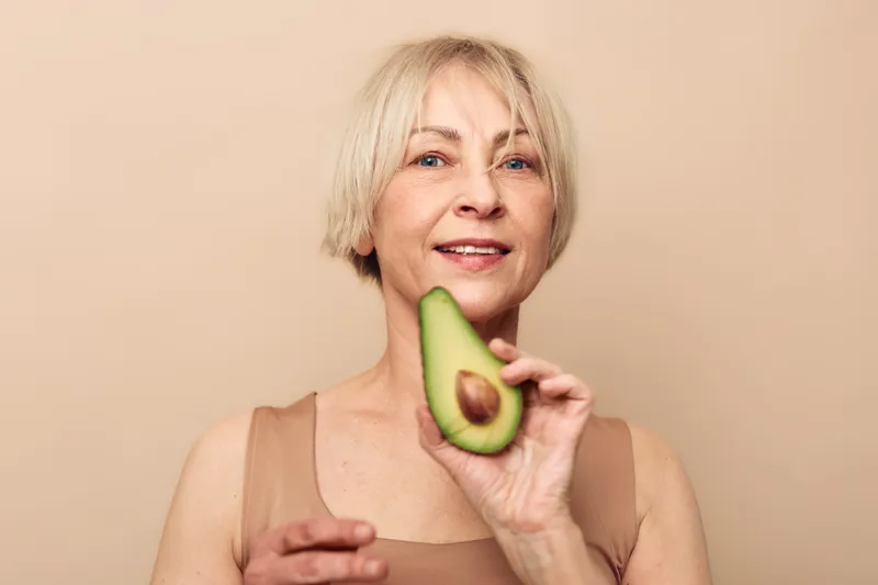 Smiling mature woman with short blonde hair holds half of fresh avocado near face on beige background. Concept of healthy eating, nutrition, wellness, and natural lifestyle for seniors.