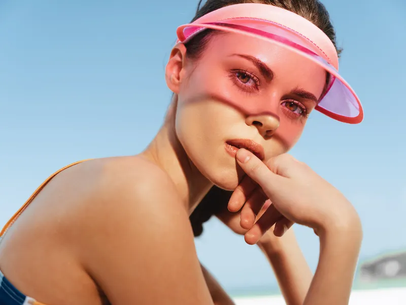 Close-up of a sunlit woman at the beach wearing a pink visor, displaying rosy skin, subtle sunburn, and a thoughtful pose that highlights summer calm and relaxed, sun-kissed mood.