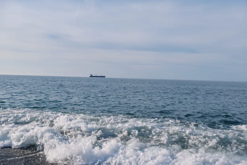 Sea view with gentle waves rolling onto a quiet shore and a distant cargo ship on the horizon, capturing vast water, blue tones and maritime calm.