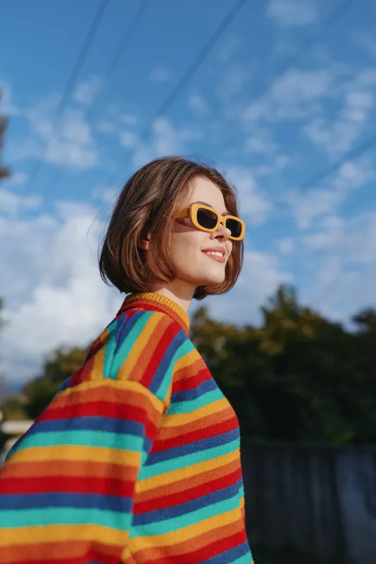 Woman in a rainbow sweater and yellow sunglasses smiling, young adult with bob haircut outdoors under sunny blue sky; casual fashion portrait with cheerful expression and travel lifestyle vibe.