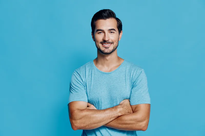 Happy smiling man with crossed arms wearing a light blue t-shirt stands confidently against a vibrant blue background. Portrait highlighting joy and self-assurance.