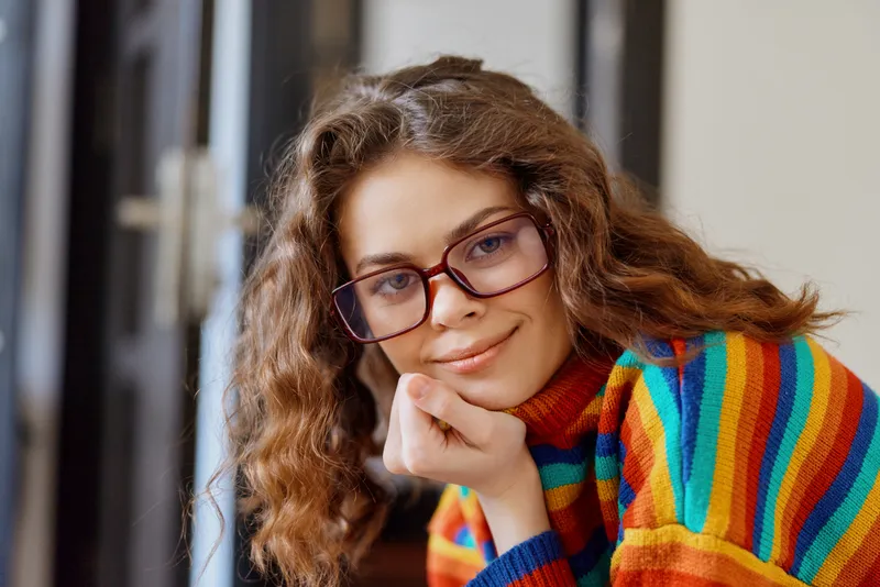Young woman with curly hair wears a colorful striped sweater and red glasses, resting her chin on her hand beside a window in a cozy home setting. The soft lighting adds warmth to mood.