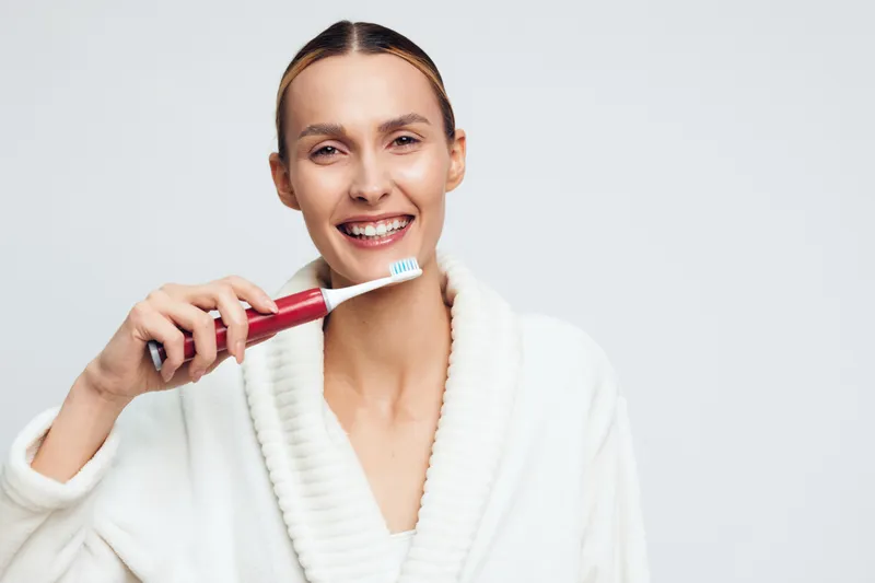 A smiling young woman in a white bathrobe brushes her teeth with a red toothbrush, standing against a pale studio background to convey a fresh morning routine.