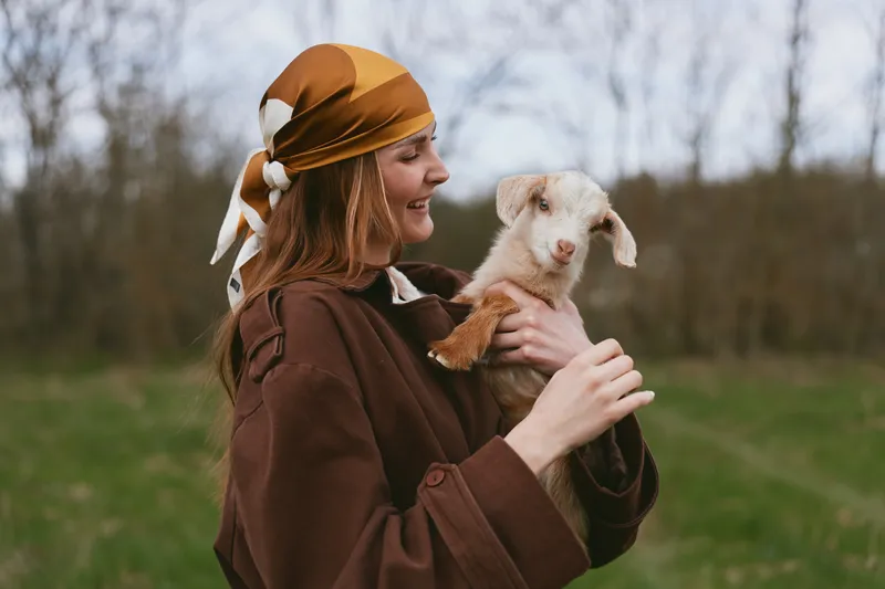 Smiling woman wearing a brown cloak holds a playful goat kid outdoors, bright natural light, rustic countryside backdrop, showcasing lifestyle, animal bond, and gentle care.