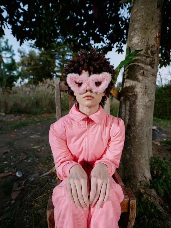 Child in a bright pink outfit seated outdoors, wearing a soft heart mask, facing the camera with a curious expression, natural background and trees creating a whimsical portrait in a park setting.