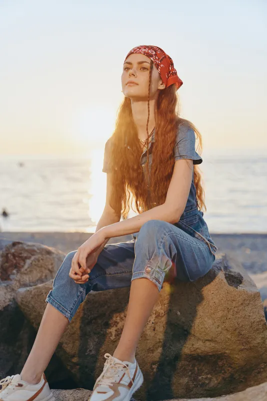 Lifestyle woman in modern boho-western denim street style with film color effect sitting on rocks by the beach at sunset, relaxed and thoughtful young woman.