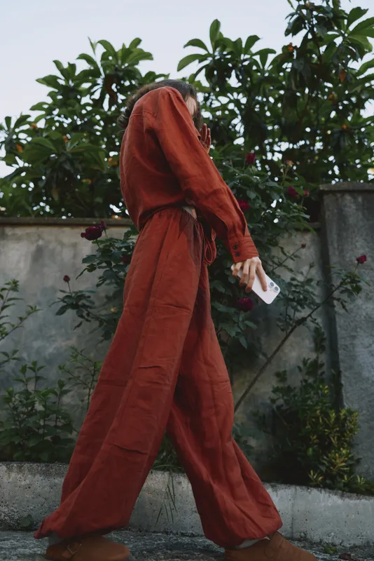 A woman in a rust red hooded jumpsuit walks along a garden path, holding a card, capturing a candid street style moment in everyday life