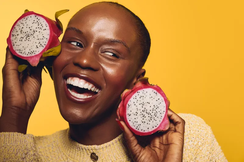 Happy African woman with dark skin holding two halves of dragon fruit near face on yellow background. Concept of healthy lifestyle, natural food, and joyful mood in studio.