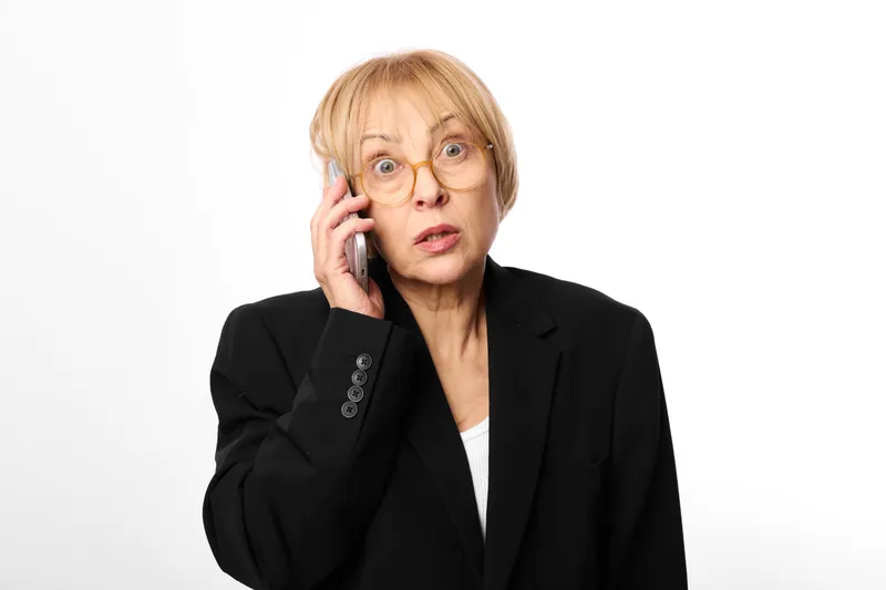 Mature woman in business suit holding a phone to her ear, surprised expression, studio portrait on white background, communication and customer service concept