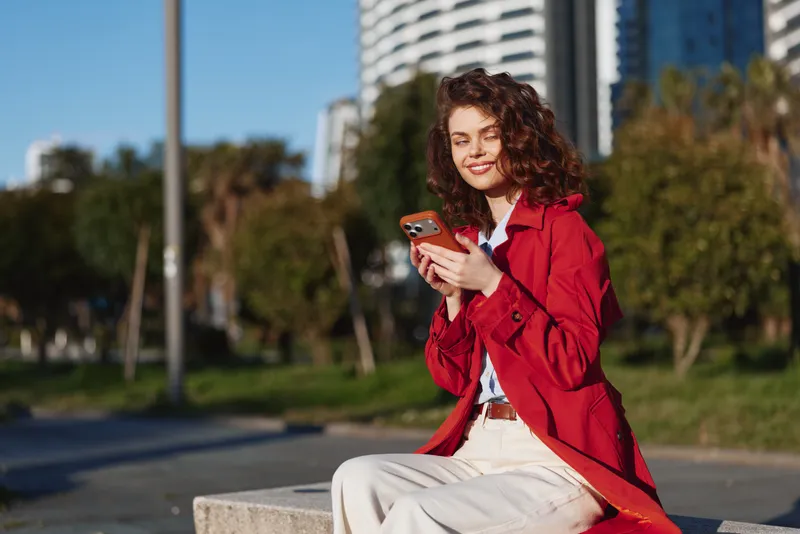Young woman in red coat sits outdoors on concrete bench uses smartphone smiles enjoys sunny day in urban park with green trees and high buildings in background.