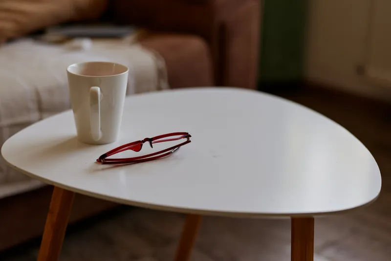 Cozy indoor living room scene features a white round table with a coffee mug and red glasses on its surface, soft natural lighting, warm tones, and a comfortable couch in the background
