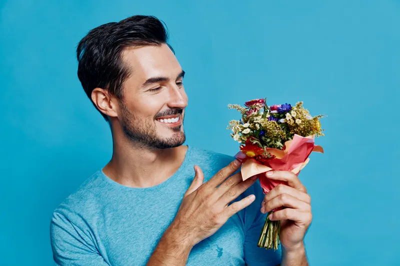 Smiling man holding a beautiful bouquet of flowers, wearing a blue shirt against a vibrant blue background, exuding happiness and warmth in studio portrait.