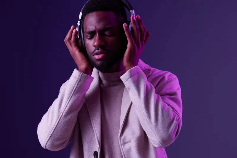 A young Black man enjoying music with headphones on, dressed in a stylish beige coat, against a vibrant purple background, conveying deep emotions and relaxation