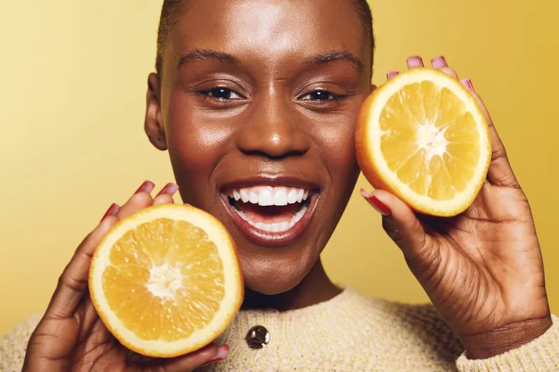 Cheerful black woman with dark skin holds two orange halves near face smiling broadly on yellow background. Concept of healthy lifestyle, vitamin C, nutrition and joyful mood.