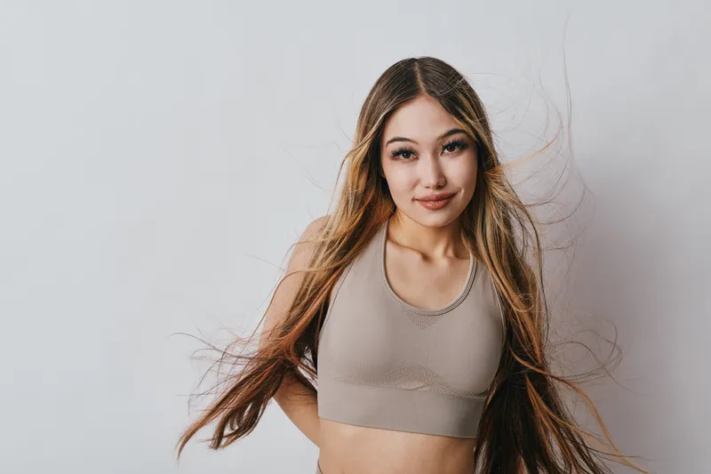 Young woman with long hair wearing beige top posing with confident smile and wind blowing through hair on light background for portrait and beauty.
