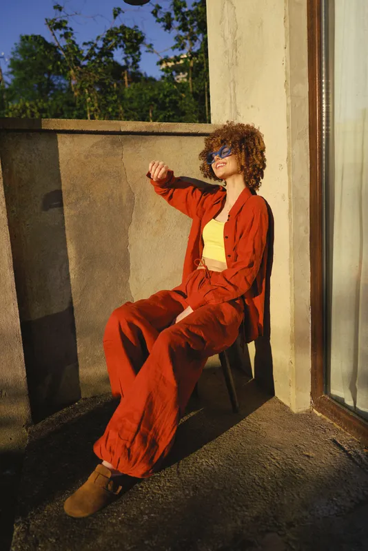 Cheerful woman outdoors, relaxed on a balcony in a red suit and yellow top, sunglasses, sunlit moment, gentle breeze, vibrant colors, urban setting