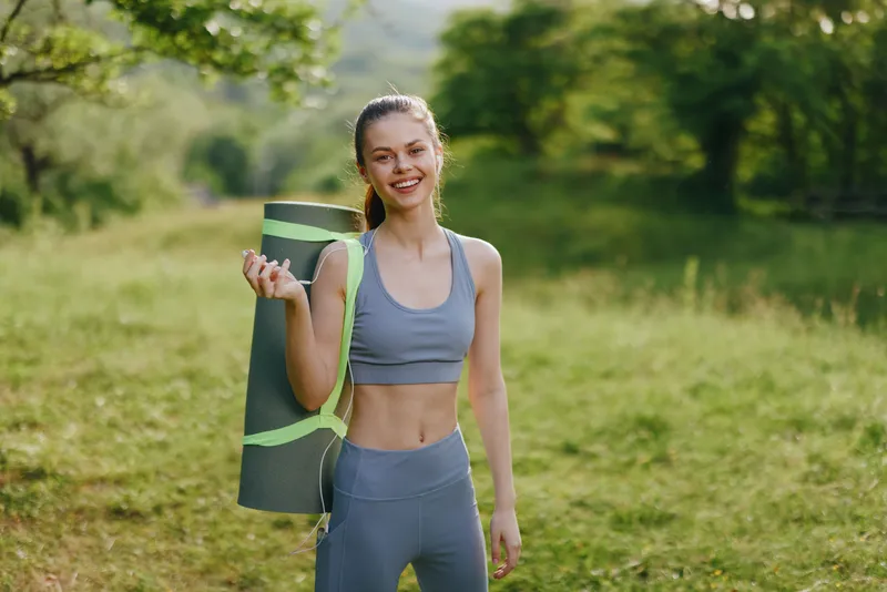 Young athletic woman in activewear holding a yoga mat outdoors, smiling happily amidst natural greenery, reflecting a vibrant and healthy lifestyle