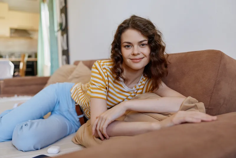 A relaxed woman lounges on a couch at home, smiling softly in a casual striped shirt. A warm living room setting conveys comfortable, everyday lifestyle and self care.