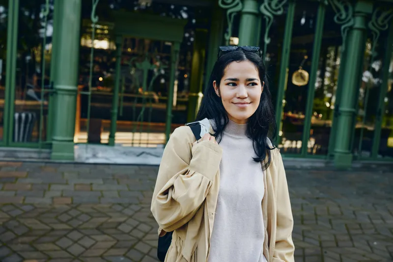 Woman in a beige jacket and gray sweater stands on a city street with a backpack, smiling softly, candid urban fashion and travel vibe for casual street style photography