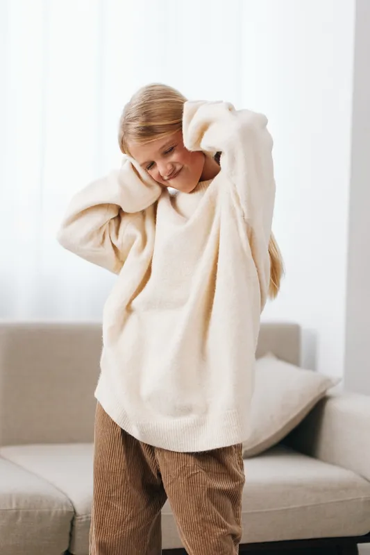 Young girl in an oversized cream sweater and corduroy pants stands in a bright living room, adjusting sleeves for a cozy, relaxed pose.