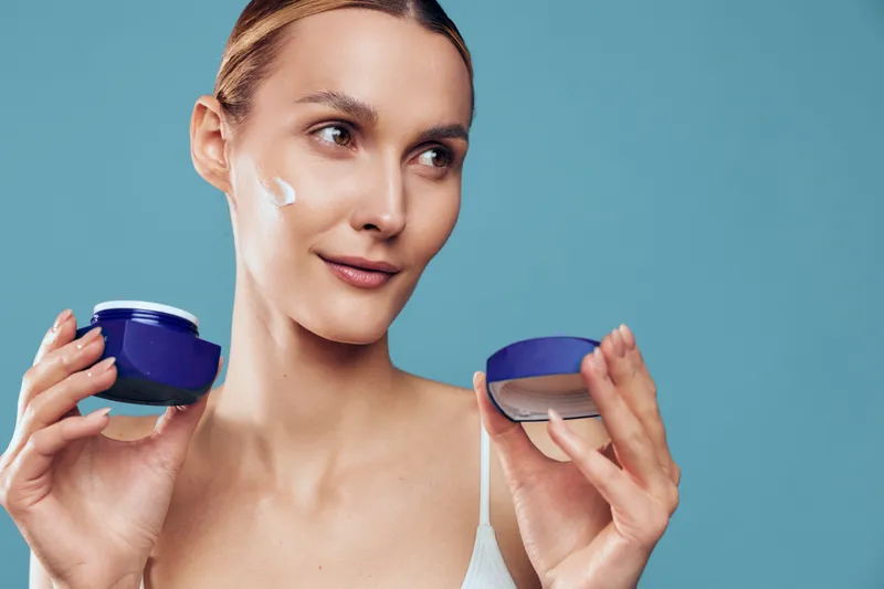 Young woman applying cream on face, holding open jar with cosmetic product, clear skin, soft natural light, blue background, skincare routine, beauty care concept.