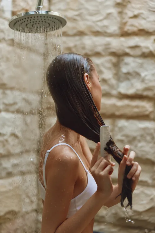 Woman combing wet hair under outdoor shower, natural light, stone wall background, casual white top, serene moment, personal care routine.