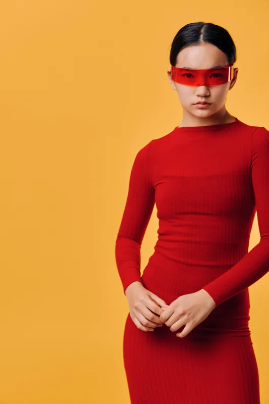Woman in red dress with long sleeves wearing red transparent glasses standing against orange background looking serious and confident with hands clasped in front.