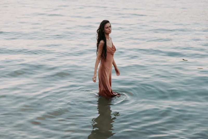 A solitary woman stands in shallow sea water, wearing a flowing peach dress, gentle waves lap around her legs, soft light creates a serene, reflective mood.