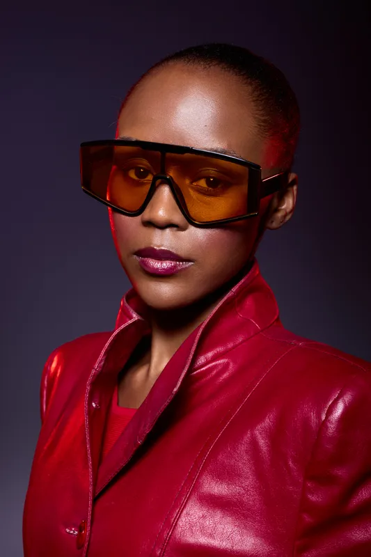 Stylish African woman with dark skin wearing oversized orange sunglasses and a red leather jacket. Studio portrait with dramatic lighting and confident expression. Fashion concept.