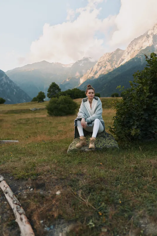 Young woman with a light jacket and hiking boots sits on a rock in a mountain meadow surrounded by greenery and distant peaks under a cloudy sky.