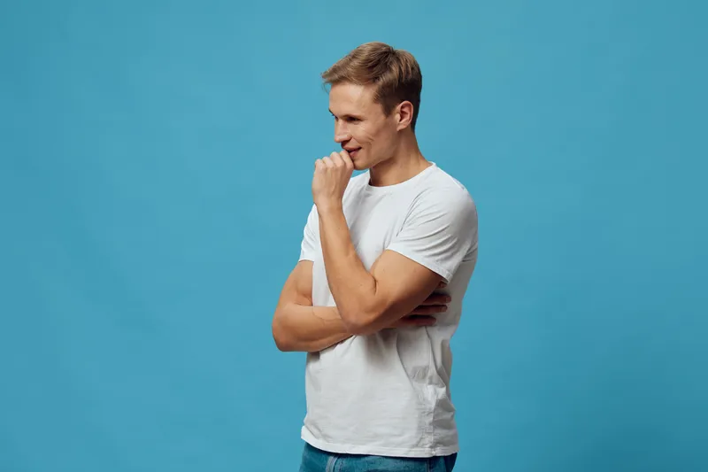 Smiling young man with light brown hair wearing casual white t-shirt and jeans, standing against a plain blue background, looking down thoughtfully with a gentle smile, indoor studio portrait. People 