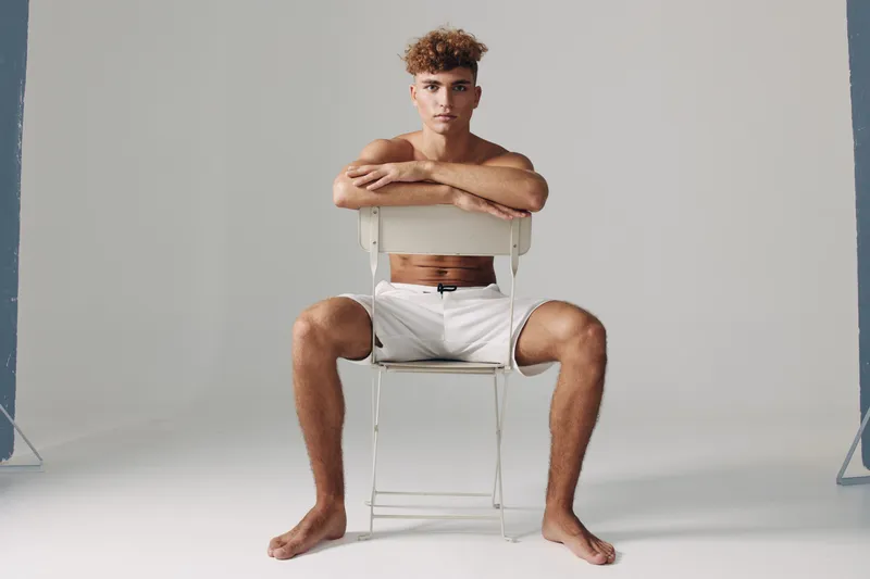 Young man sitting barefoot on chair backwards with arms resting on backrest in studio. Casual pose with white shorts and neutral background for fitness or lifestyle concept.
