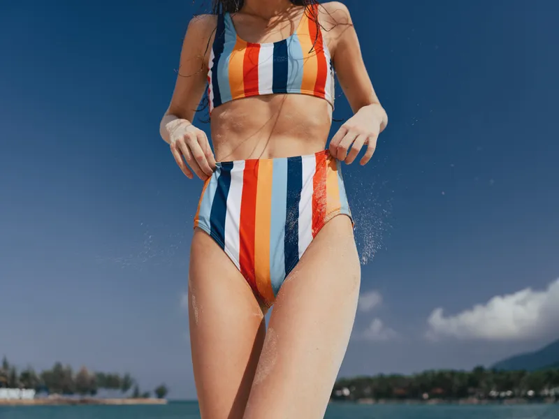 Close up of a person wearing a colorful striped bikini standing on a sunny beach with a clear blue sky, spray of water, and a carefree summer vibe.