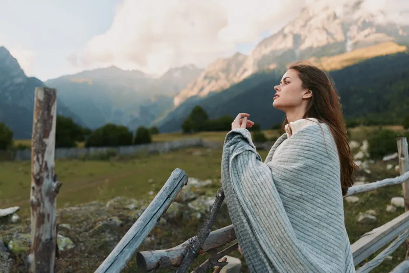 Woman wrapped in a gray knit blanket stands outdoors, gazing upward with mountains in the background, evoking a cozy, serene lifestyle moment in nature.