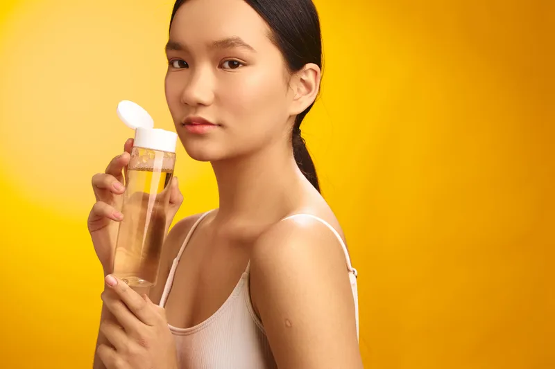 Asian woman holds a bottle of lotion against a warm yellow background, highlighting skincare and beauty. She looks at the camera with calm confidence, creating a fresh, inviting lifestyle moment