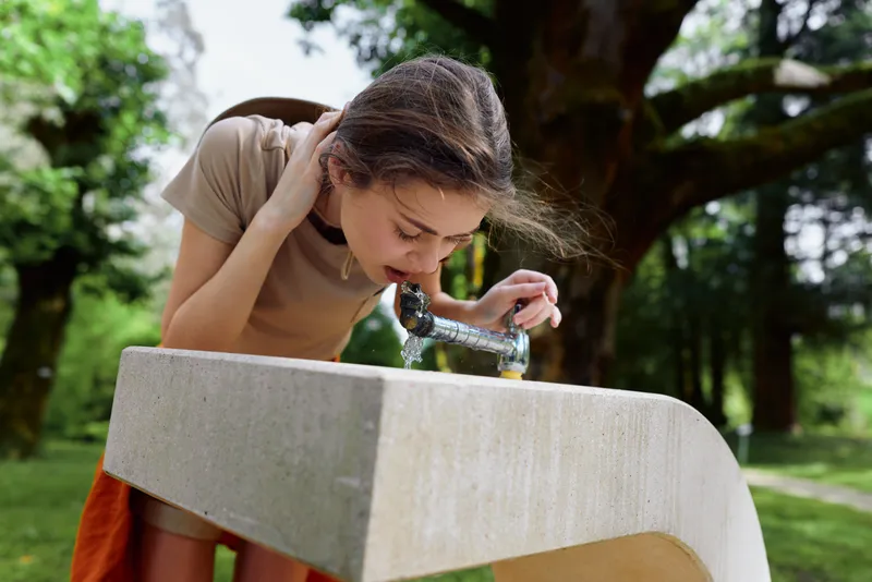 Girl drinking water from outdoor fountain in park, child leaning over concrete basin to sip from metal spout on a summer day, casual clothing, green trees and sunlight in background