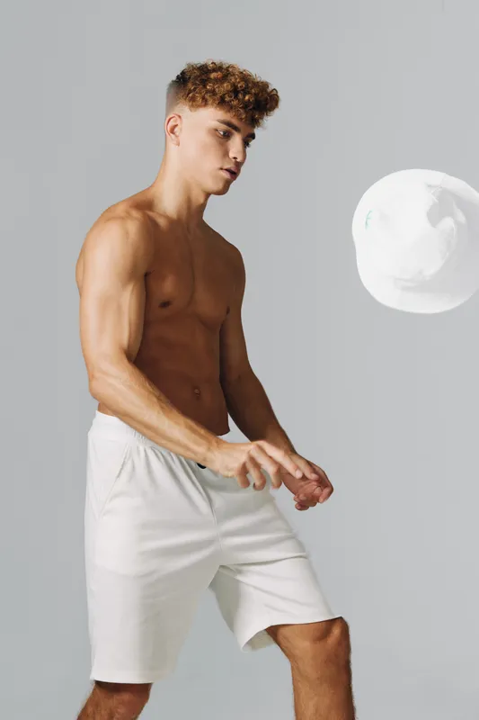 Young athletic man with curly hair wearing white shorts playing with a ball, posing on grey background in studio for sports, fitness, activity, motion and exercise concept with healthy lifestyle.