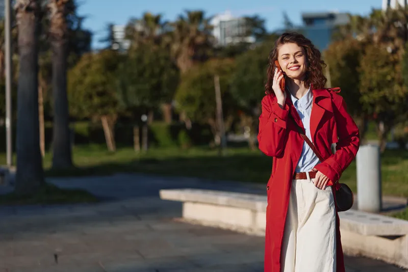 Smiling young woman with curly hair wearing a red coat stands outdoors in a sunny park, talking on a phone as casual city life unfolds nearby.