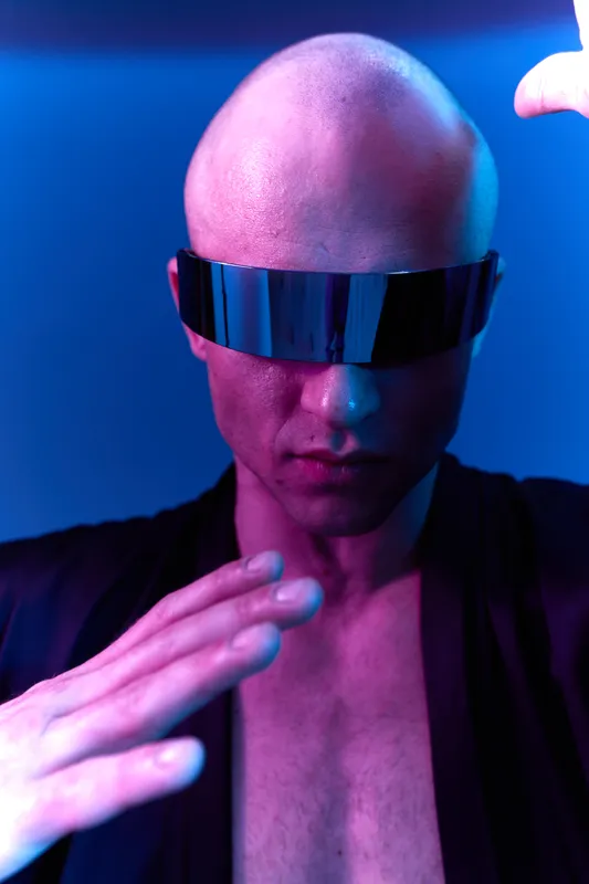 A striking man with a bald head and futuristic sunglasses showcases a captivating pose, evoking a sense of mystery and intrigue in vibrant color light.