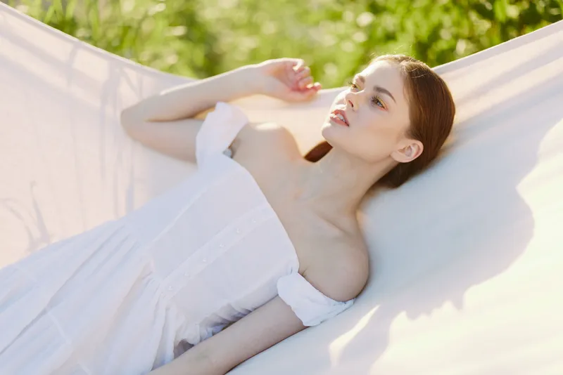 Young woman in a white dress relaxing in a hammock, enjoying a sunny day with a serene expression amidst nature s green background