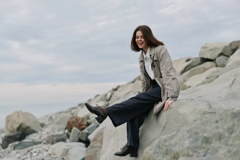 Young woman in casual clothes sits on large rocks by the sea, smiling and enjoying the outdoors. Cloudy sky and rocky beach create a relaxed natural atmosphere.