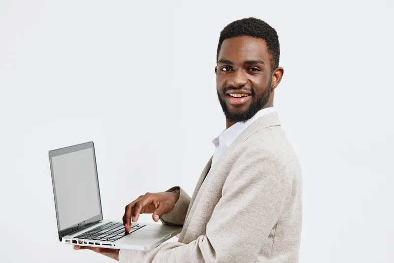 Smiling young African American man in cozy light beige sweater working on silver laptop isolated on plain white background, studio portrait. People lifestyle concept, technology, casual fashion, posit