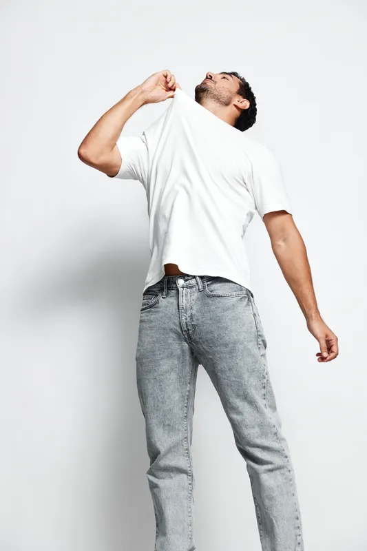Full body portrait of a stylish young man in a white t-shirt and gray jeans, playfully pulling his shirt while looking upwards against a solid neutral background. Fashion concept.