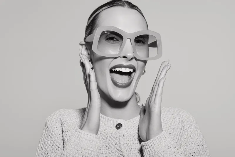 Happy white woman with light skin wearing oversized sunglasses and knitted sweater in studio. Concept of fashion, joy, and modern style.