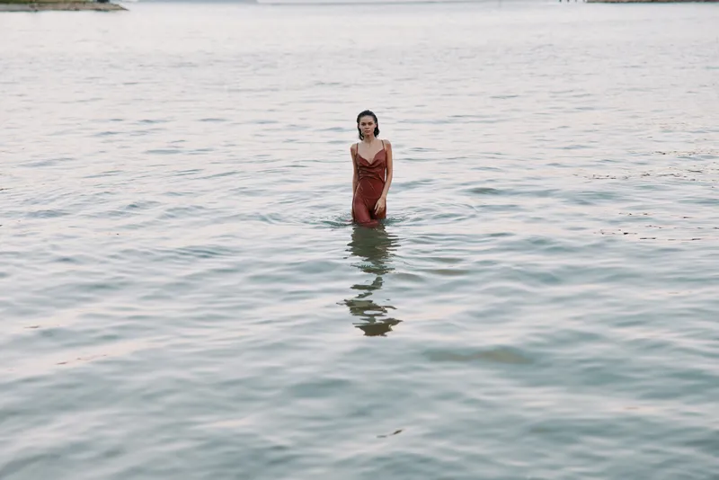 Woman stands waist deep in clear sea water wearing a flowing red dress, gentle breeze tousling hair, serene scene highlighting solitude, beauty and muted shoreline tones.